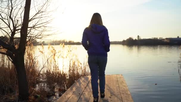 一个穿着黑色运动鞋 蓝色夹克和牛仔裤的女孩 女人沿着木码头散步 走到边缘 在初春或初秋的日落时停止在池塘边射击 — 图库视频影像