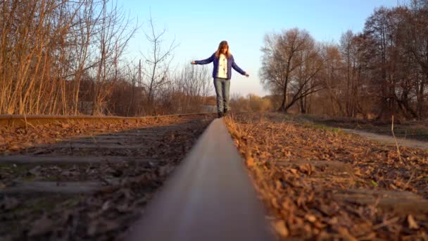 青少年穿着黑色运动鞋 蓝色夹克 白色毛衣和牛仔裤走在铁轨上走向相机 拍摄在春天 秋天的春天 秋天的日落 — 图库视频影像