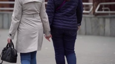 İki kız: biri kot pantolon ve lacivert ceket, diğeri kot pantolon ve açık pelerinli, el ele tutuşarak sokakta yürürken, kadın arkadaşlığı, eşcinsel aşk, arkadan ateş etmek.