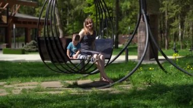 Küçük bir çocuk, bir erkek ile bir anne, güneşli bir günde ilkbahar veya yaz aylarında Park'ta siyah metal salıncak salıncak, bir kadın kameraya bakar, sonra çocuk, çocuk diyor, Park, famil dinlenme