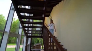 Beyaz tişörtlü bir kadın, kot pantolon ve siyah güneş gözlüğü metal merdivenlerden aşağı yürüyor, panoramik Windows ile salon, yaz Güneşli gün