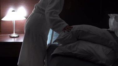 Yatak için hazırlamak için beyaz bir Terry Robe bir kadın: yatak yaklaşımlar, battaniye kaldırır, yastıklar yatak, yapay aydınlatma ve loş ışık, ortalama planı battaniye kaldırır ayarlar