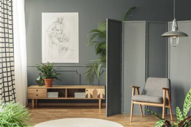 Retro armchair against a gray screen next to a drawing hanging on a dark wall with molding above wooden cupboard in living room interior clipart