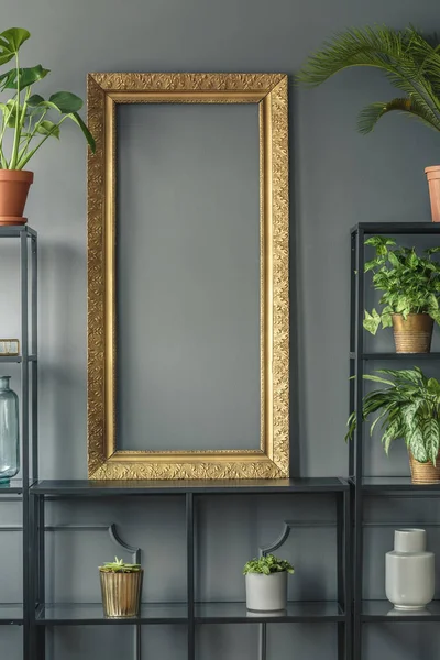 A gold frame and plants in vases on black shelves next to a grey wall