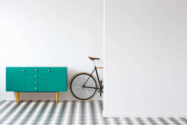 Green cabinet next to a bike against white wall in flat interior with checkerboard floor and copy space