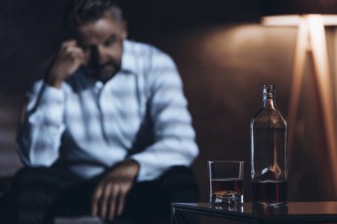 Karanlık bir odada şişe ve cam alkol ile oturan adam vurguladı