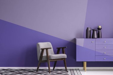 Real photo of a gray, wooden armchair on patterned, black and white rug in creative living room interior with geometric, violet wall and cupboard clipart