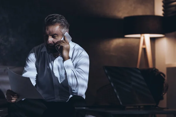 Mężczyzna Rozmawia Przez Telefon Czytanie Dokumentów Ciemnym Pomieszczeniu Średnim Wieku — Zdjęcie stockowe