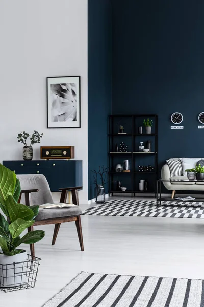 Dark living room interior with armchair, plant, striped rugs, metal shelf and sofa in the background