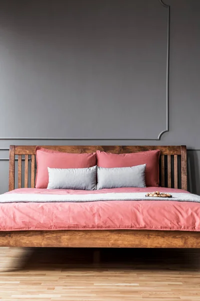 Close-up of a simple bed with wooden bedhead and pink sheets in dark, elegant bedroom interior with molding on dark walls. Real photo