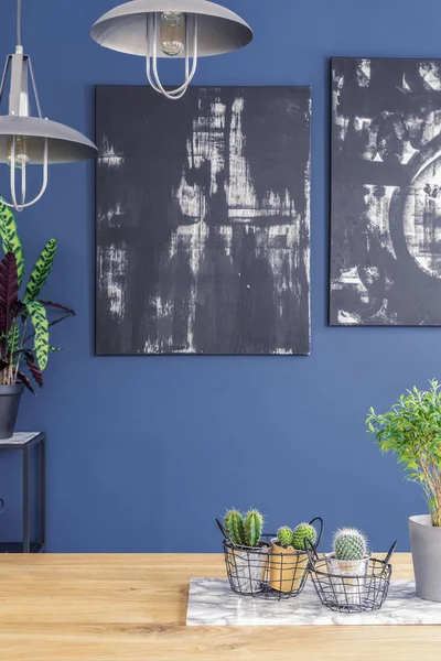 Black paintings on a blue wall and industrial pendant lights over a wooden kitchen interior table with plants and a marble board