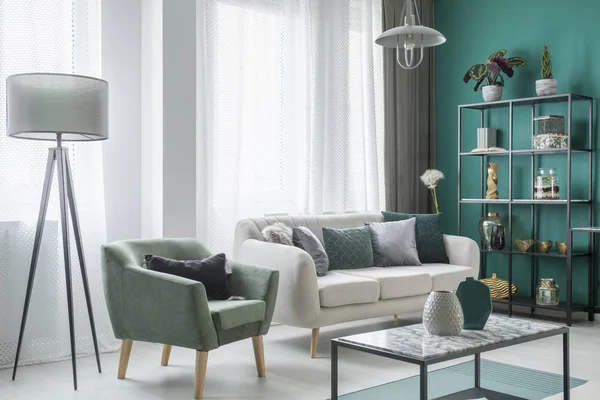 Lamp next to a green armchair and beige sofa in living room interior with decoration on table