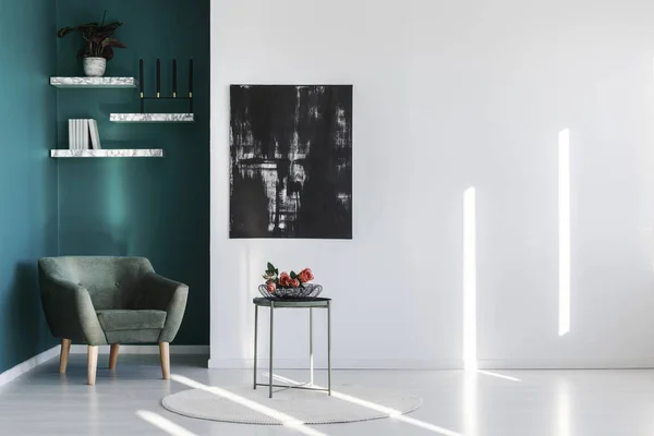 Comfortable, dark green armchair in a reading corner of a minimalist, white and teal living room interior