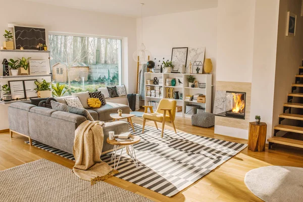 Blanket thrown on grey corner sofa in white living room interior with fresh plants, carpet, fireplace and simple posters