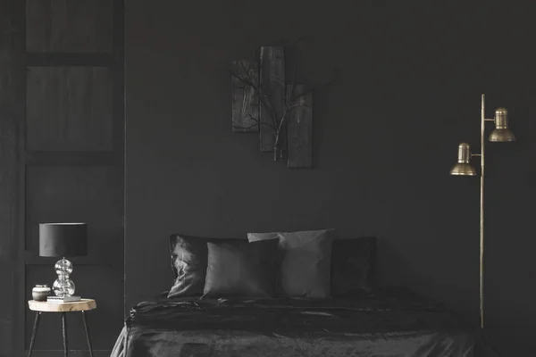 Gold lamp next to bed in black bedroom interior with sculpture on dark wall