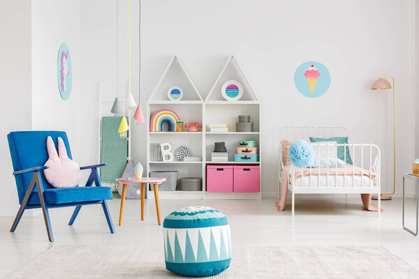 Patterned pouf near blue armchair in colorful kid's room interior with a poster above white bed