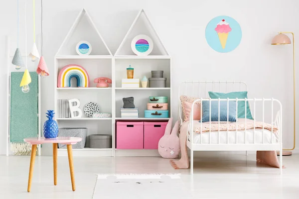 Front view of a kid\'s room interior with a table, shelves with boxes and rainbow, single bed and ice-cream poster on the wall