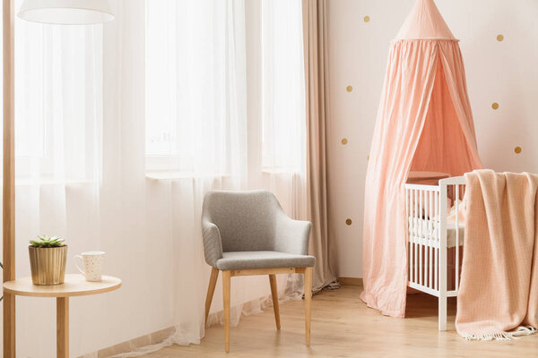 Wooden crib with dirty pink canopy standing in bright nursery room interior with grey chair and windows