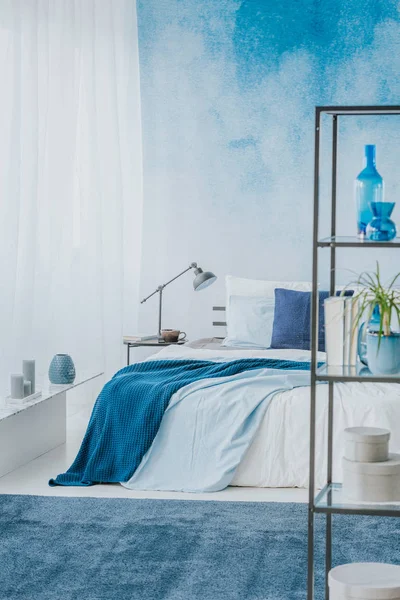 Double bed with blue blankets and black, metal shelf in a sky blue bedroom interior