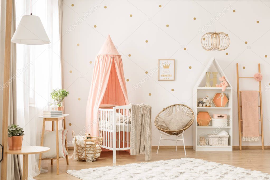 Gold chair with pillow standing next to a white wooden crib with pastel pink canopy in bright baby room interior with dots on the wall