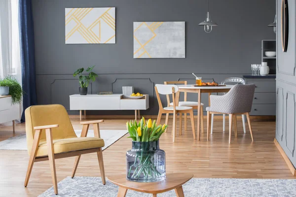 としたリビング ルームのインテリアの肘掛け椅子の横にあるテーブルの上の灰色の壁に黄色の絵画とチューリップ — ストック写真