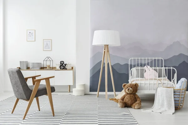 A wooden floor lamp and a modern gray armchair in a monochromatic child\'s bedroom interior in scandinavian style