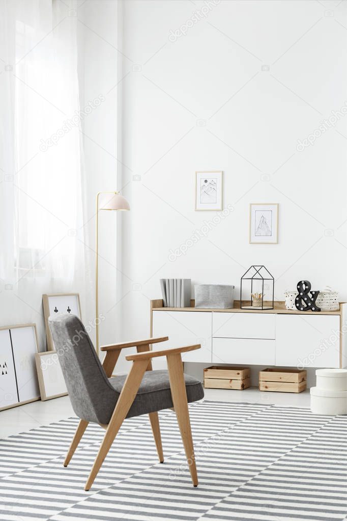 Wooden crates under a white sideboard and a comfortable, modern gray armchair in a stylish living room interior with a striped rug and white wall
