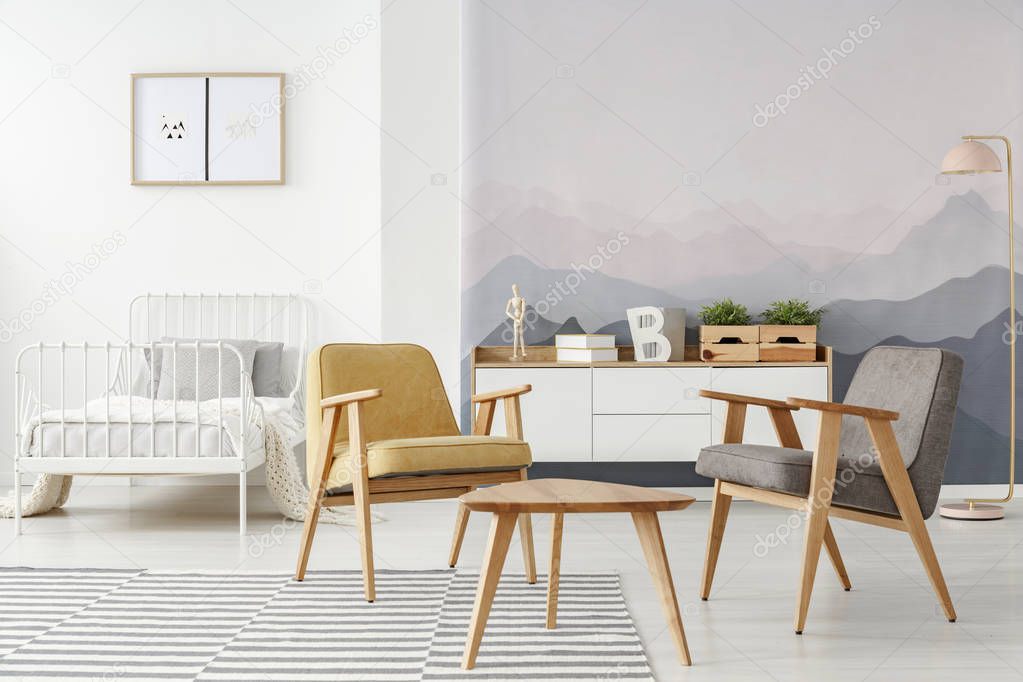 Yellow and gray modern armchairs and a white bed in a multifunctional room interior with mountains wallpaper and wooden furniture
