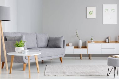 Table next to grey sofa in scandi living room interior with posters above cupboard. Real photo clipart