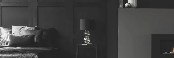 Real photo of an elegant lamp with a glass base on a simple night stand between a cozy bed and fireplace in black apartment room interior