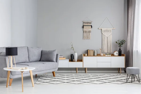 Lamp on table next to grey sofa in scandi living room interior with DIY decor above cupboard. Real photo