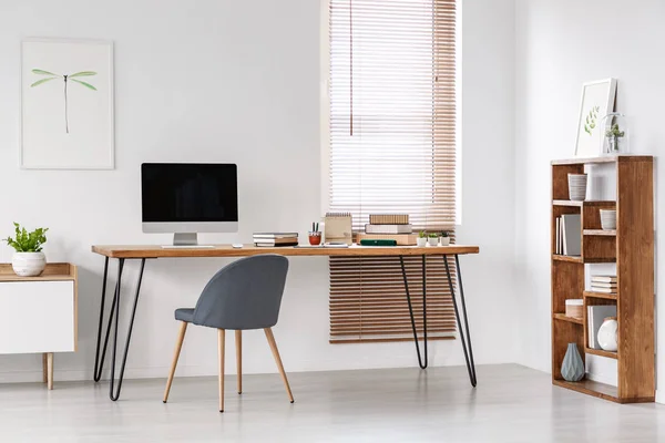 Grey chair at desk with computer monitor in minimal workspace interior with poster. Real photo