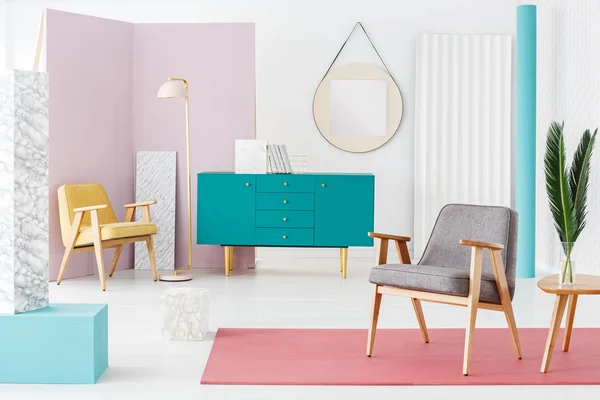 Leaves on wooden table next to a grey armchair on pink rug in flat interior with green cabinet