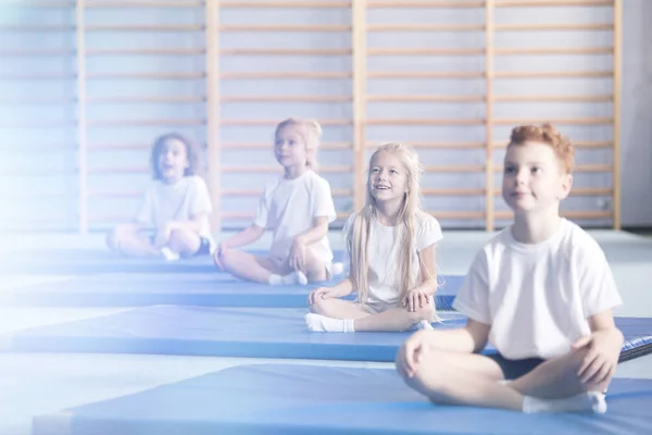 Curious and amazed children in school sportswear sitting in a gym interior during extracurricular yoga class and looking up in the direction of a flare