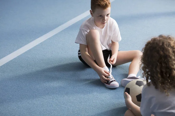 少年サッカーを開催し スポーツの靴を結ぶこと友人の向かい青いジムの床の上に座っての肖像画 — ストック写真