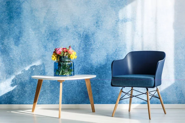 シンプルなリビング ルームのインテリアの海軍青い肘掛け椅子の横にある白い木製テーブルに色とりどりの花 — ストック写真