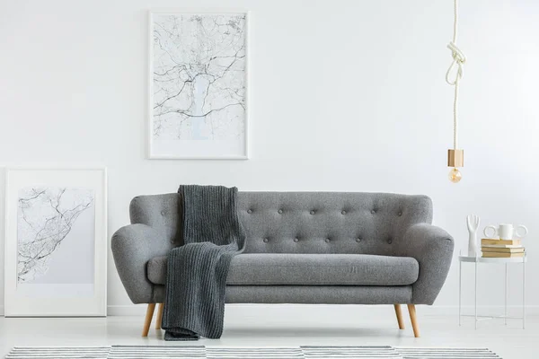 Grey lounge with dark blanket standing in white living room interior with two map posters, gold bulb lamp and books on a small table