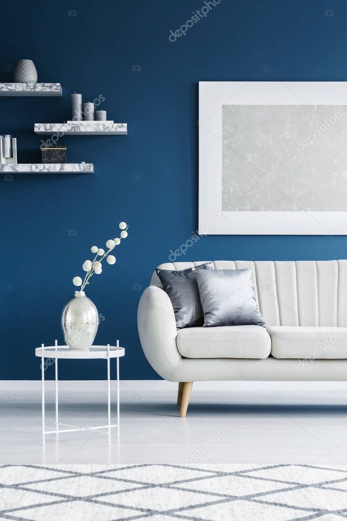 White, leather couch with silver pillows next to a side table with a vase set on a blue wall