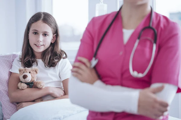 在诊所逗留期间 微笑的女孩与长毛绒玩具 模糊的照顾者穿着粉红色制服的前景 — 图库照片