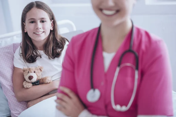 微笑生病的女孩与长毛绒玩具 医生在粉红色制服的前景 — 图库照片