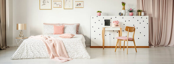 Bedside table with glass lamp standing next to king-size bed with dotted sheets, pink pillows and blanket standing in white bedroom interior with posters, fresh plants and dressing table with telephone