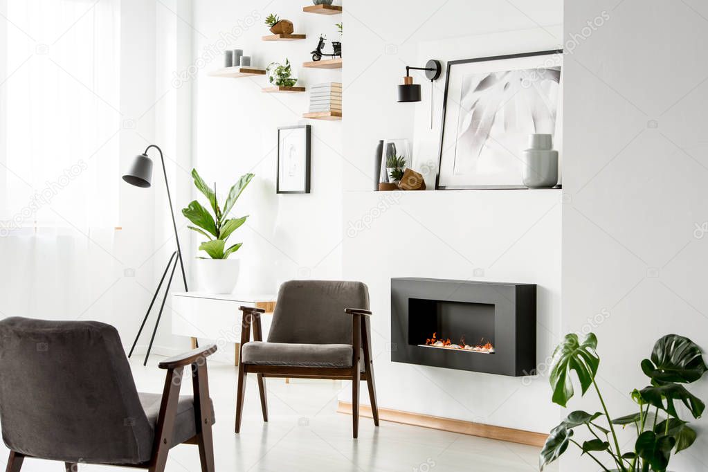 Grey armchairs next to fireplace in white apartment interior with plant, poster and lamp. Real photo