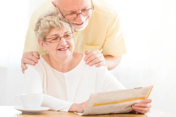 年配の女性が読む新聞と彼女の夫は彼女を抱き締める朝の笑顔 — ストック写真