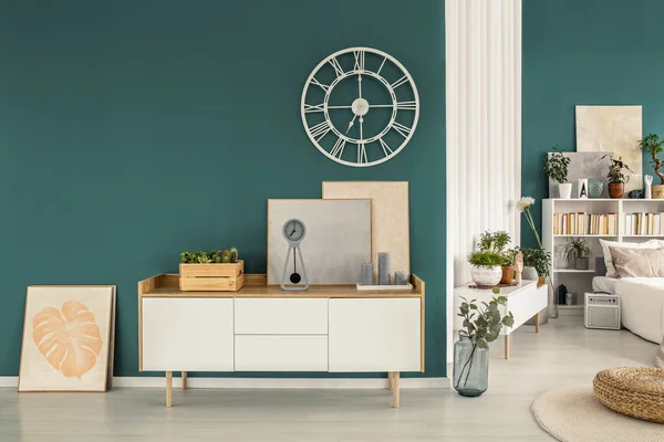 White cabinet, paintings and modern clock on a green wall with a bedroom interior in the background