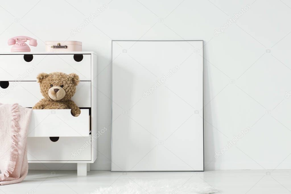 Plush toy and pink blanket in cabinet next to empty white poster in baby's room interior. Real photo. Paste your photo here