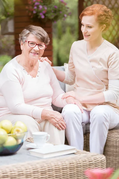 穿着制服的专业护理人员把她的手放在一个老太太的肩膀上 在一个老人家的院子里吃零食 背景模糊 — 图库照片