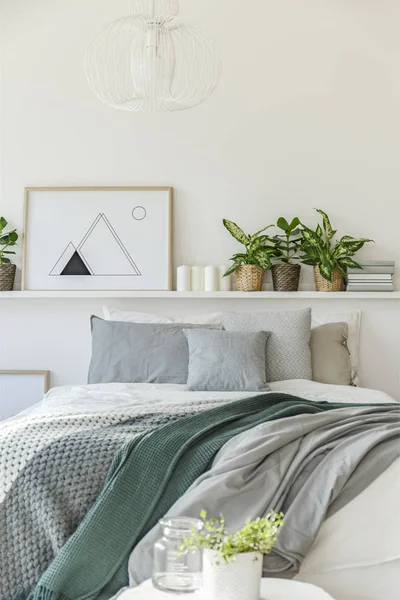 Grey and green blanket on bed against the wall with poster, plants and candles in bedroom interior