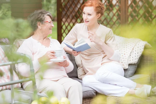 Caregiver Membaca Buku Untuk Seorang Wanita Tua Minum Teh Teras — Stok Foto