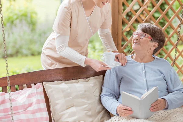 Nurse giving tea to happy elderly woman reading book on the terrace