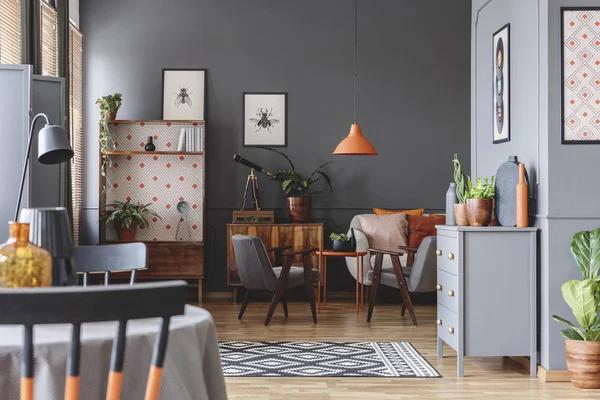 Orange lamp above table and grey armchair in retro living room interior with telescope on cabinet